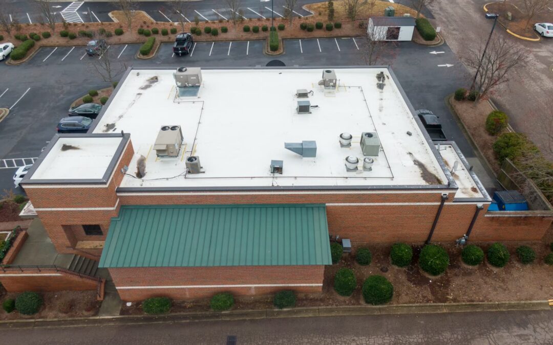 Aerial view of a brick commercial building with a white TPO flat roof, rooftop HVAC units and a green standing-seam metal roof section